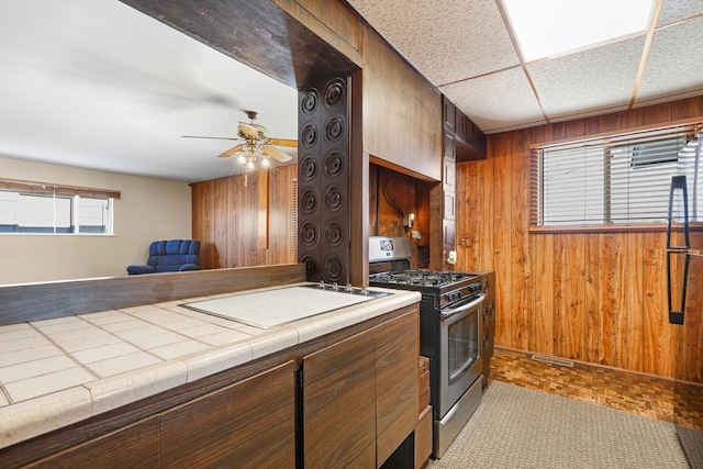 kitchen featuring stainless steel range with gas cooktop, tile counters, a drop ceiling, wooden walls, and ceiling fan