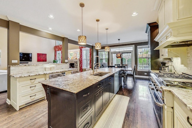 kitchen featuring custom range hood, an island with sink, hanging light fixtures, light stone countertops, and appliances with stainless steel finishes