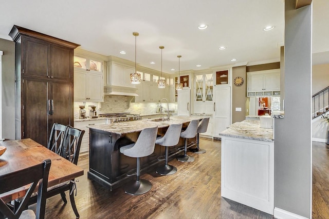 kitchen featuring a kitchen island with sink, pendant lighting, light stone counters, and dark wood-type flooring