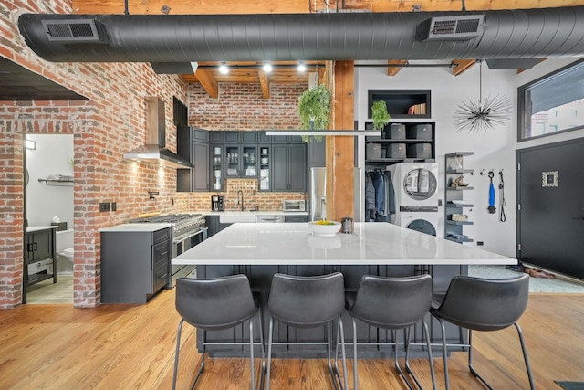 kitchen featuring light wood-type flooring, stainless steel stove, wall chimney exhaust hood, and brick wall