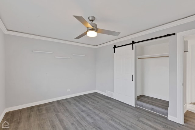 unfurnished bedroom featuring ceiling fan, a barn door, crown molding, wood-type flooring, and a closet