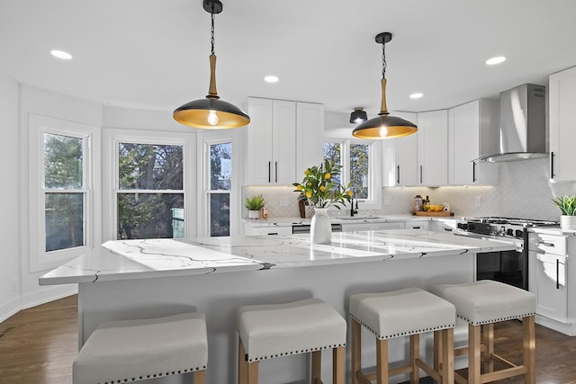 kitchen with wall chimney exhaust hood, light stone countertops, gas range oven, a kitchen island, and white cabinetry