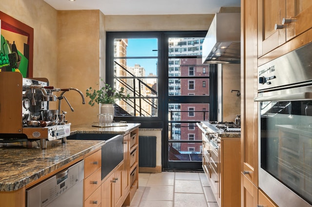 kitchen with appliances with stainless steel finishes, wall chimney exhaust hood, sink, light tile patterned floors, and dark stone countertops