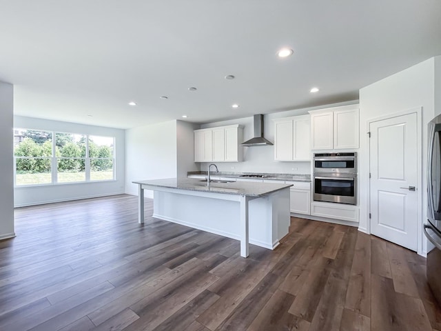 kitchen featuring light stone countertops, an island with sink, white cabinetry, and wall chimney range hood