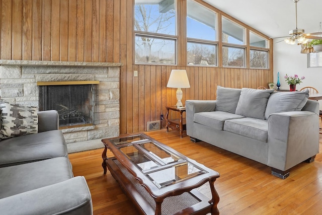 living room featuring ceiling fan, wooden walls, wood-type flooring, a stone fireplace, and lofted ceiling