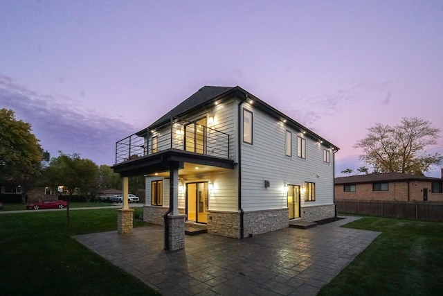 back house at dusk with a lawn, a balcony, and a patio