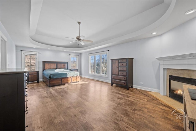 bedroom featuring ceiling fan, a tiled fireplace, a tray ceiling, and dark hardwood / wood-style flooring