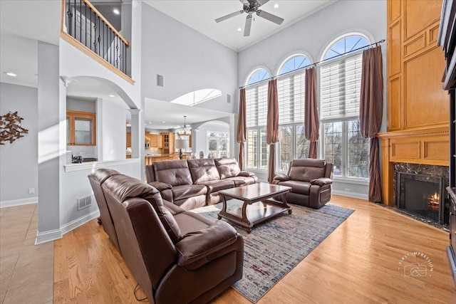 living room featuring a high end fireplace, light hardwood / wood-style floors, a high ceiling, decorative columns, and ceiling fan with notable chandelier