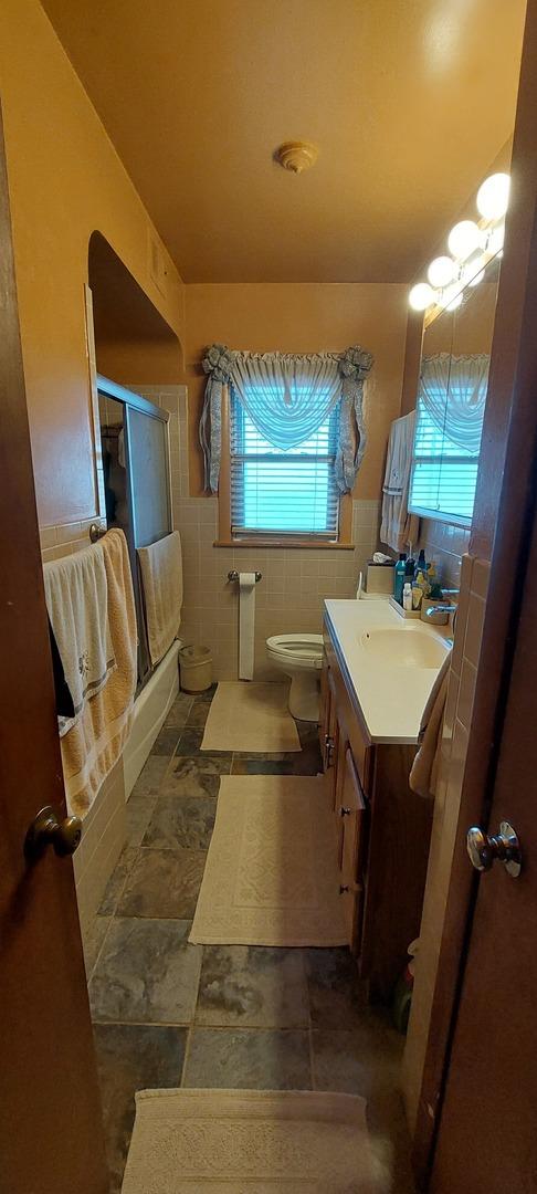 full bathroom with combined bath / shower with glass door, toilet, tile walls, and vanity