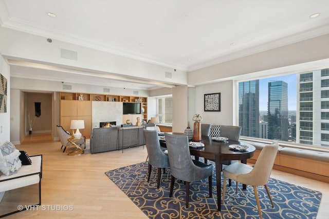 dining area with light wood-type flooring, a warm lit fireplace, crown molding, and recessed lighting