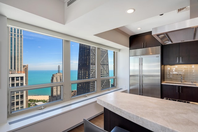 kitchen featuring dark brown cabinetry, sink, tasteful backsplash, stainless steel built in fridge, and a water view