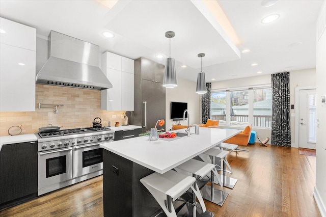 kitchen with wall chimney exhaust hood, range with two ovens, white cabinets, hanging light fixtures, and an island with sink