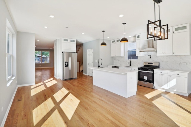 kitchen with pendant lighting, wall chimney range hood, an island with sink, appliances with stainless steel finishes, and white cabinetry