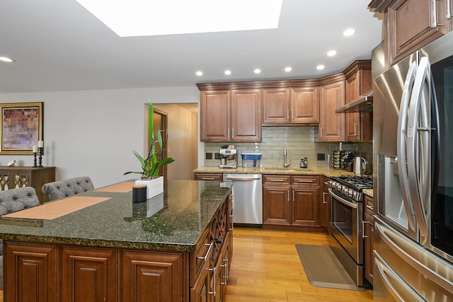 kitchen with appliances with stainless steel finishes, dark stone countertops, a skylight, a center island, and light wood-type flooring