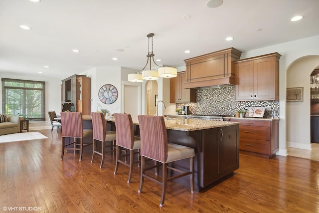 kitchen with pendant lighting, dark wood-type flooring, a kitchen island with sink, light stone countertops, and a kitchen bar