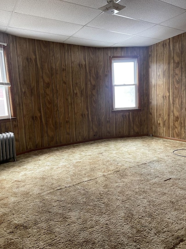 empty room with a paneled ceiling, wood walls, radiator heating unit, and carpet floors