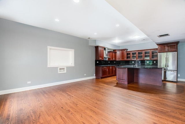 kitchen featuring light wood-type flooring, wall chimney range hood, sink, a center island with sink, and stainless steel refrigerator