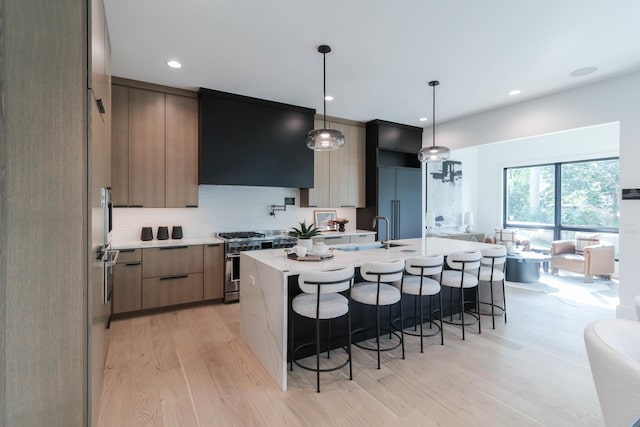 kitchen with ventilation hood, high end stainless steel range oven, a kitchen island with sink, decorative light fixtures, and light hardwood / wood-style flooring