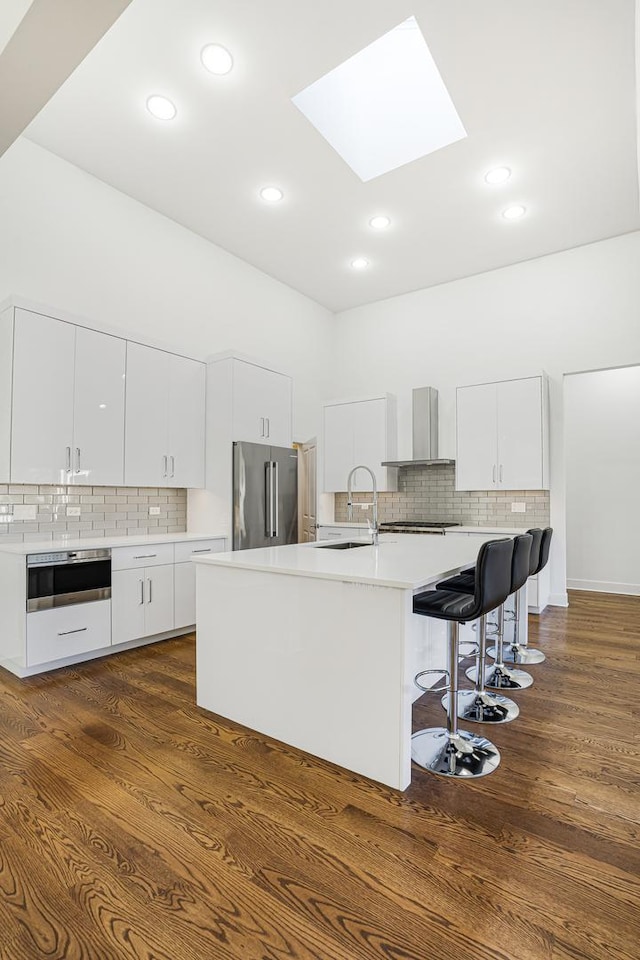 kitchen featuring a skylight, wall chimney exhaust hood, high end refrigerator, a center island with sink, and white cabinets