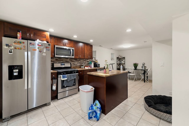 kitchen with appliances with stainless steel finishes, backsplash, light stone counters, light tile patterned floors, and a kitchen island