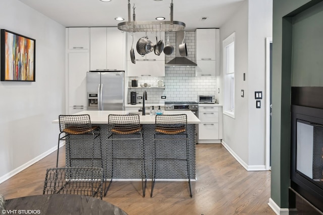 kitchen featuring wall chimney exhaust hood, backsplash, an island with sink, white cabinets, and appliances with stainless steel finishes