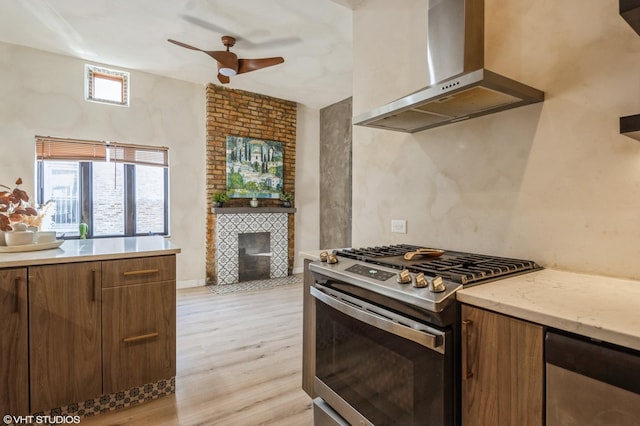 kitchen featuring light wood finished floors, a brick fireplace, stainless steel gas stove, ceiling fan, and wall chimney exhaust hood