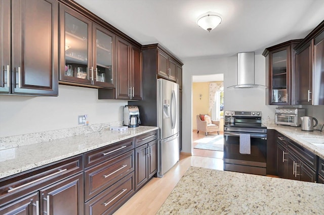 kitchen with light stone counters, wall chimney exhaust hood, dark brown cabinets, stainless steel appliances, and light hardwood / wood-style floors