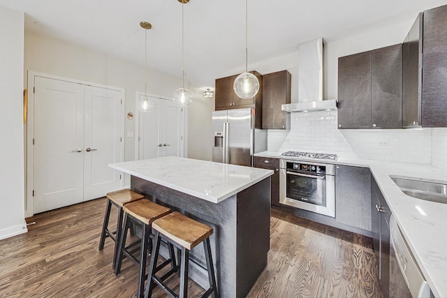 kitchen featuring stainless steel appliances, a kitchen bar, wall chimney exhaust hood, dark brown cabinets, and backsplash