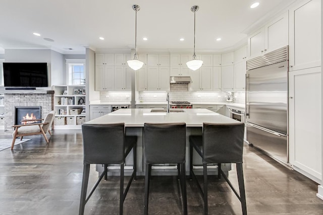 kitchen with hanging light fixtures, a center island with sink, built in fridge, white cabinetry, and sink