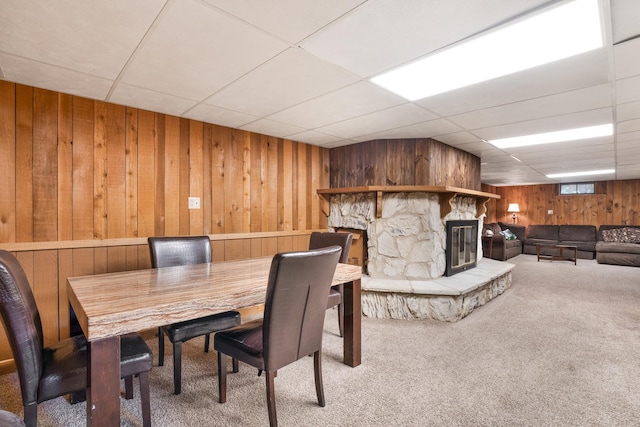 carpeted dining room with a paneled ceiling, wood walls, and a stone fireplace