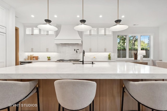 kitchen featuring pendant lighting, custom exhaust hood, a kitchen island with sink, sink, and white cabinetry