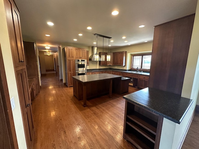 kitchen with stainless steel double oven, sink, wall chimney range hood, decorative light fixtures, and a center island