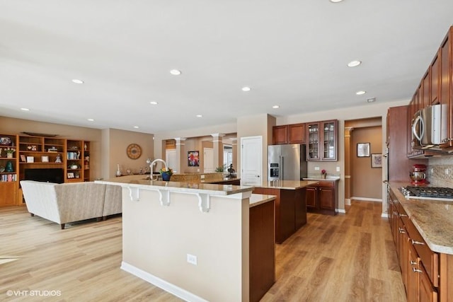 kitchen featuring light wood-style flooring, a kitchen breakfast bar, appliances with stainless steel finishes, a large island with sink, and ornate columns