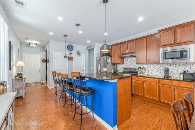 kitchen featuring wood-type flooring, hanging light fixtures, dark stone countertops, an island with sink, and stainless steel appliances