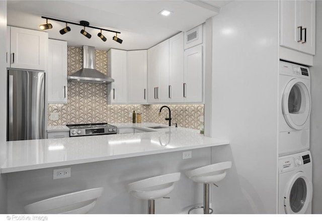 kitchen with white cabinetry, sink, stainless steel appliances, wall chimney range hood, and stacked washer and clothes dryer