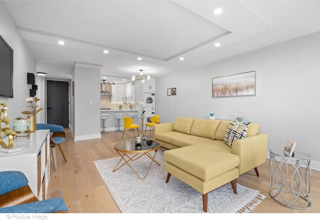 living room featuring light wood-type flooring, a tray ceiling, stacked washer / drying machine, and sink