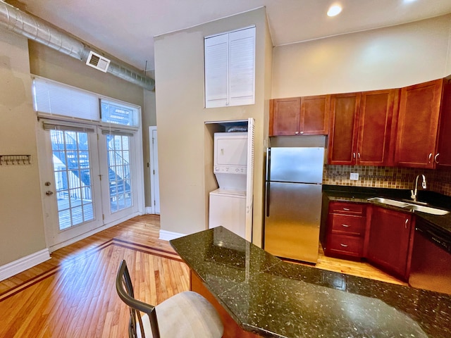 kitchen with sink, dark stone counters, stacked washer / drying machine, appliances with stainless steel finishes, and light wood-type flooring