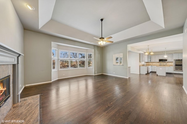 unfurnished living room with ceiling fan with notable chandelier, a raised ceiling, dark wood-type flooring, and a tiled fireplace