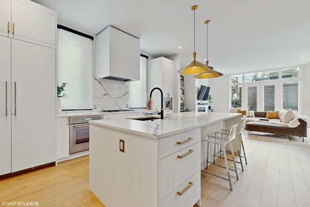 kitchen with stainless steel oven, decorative light fixtures, a center island with sink, tasteful backsplash, and white cabinetry