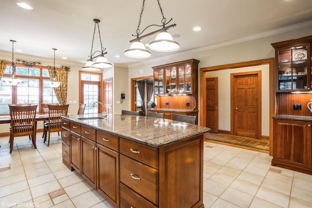 kitchen featuring sink, ornamental molding, an island with sink, dark stone counters, and hanging light fixtures