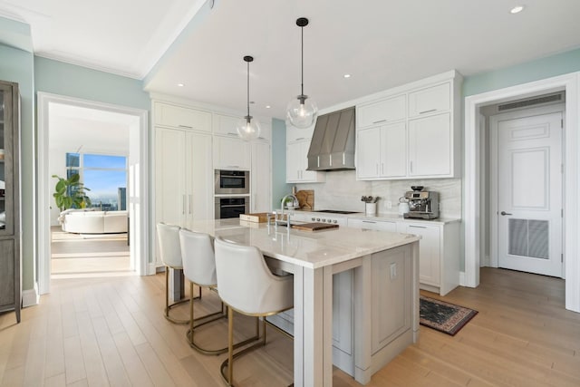 kitchen with white cabinetry, hanging light fixtures, a kitchen island with sink, wall chimney range hood, and decorative backsplash