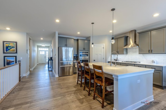 kitchen with wall chimney exhaust hood, a kitchen bar, a center island with sink, pendant lighting, and appliances with stainless steel finishes