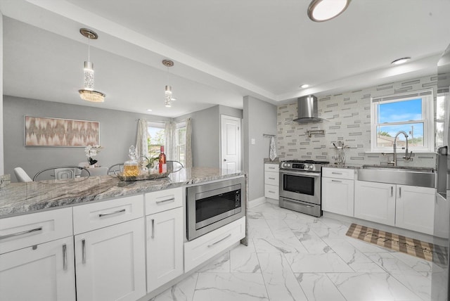 kitchen featuring sink, stainless steel appliances, wall chimney range hood, decorative light fixtures, and white cabinets