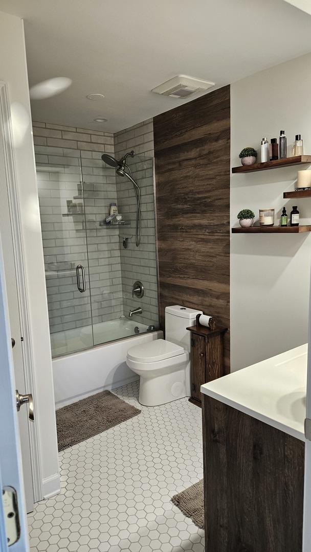 full bathroom featuring toilet, shower / bath combination with glass door, wood walls, and vanity