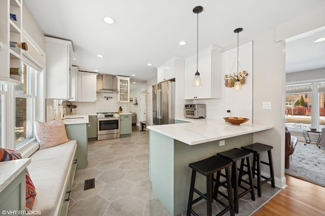 kitchen with white cabinets, appliances with stainless steel finishes, wall chimney exhaust hood, hanging light fixtures, and a breakfast bar area