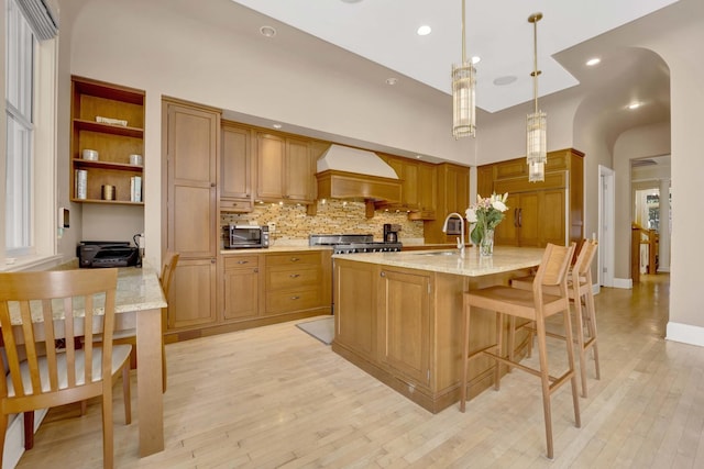 kitchen with light stone counters, stove, pendant lighting, a center island with sink, and custom exhaust hood