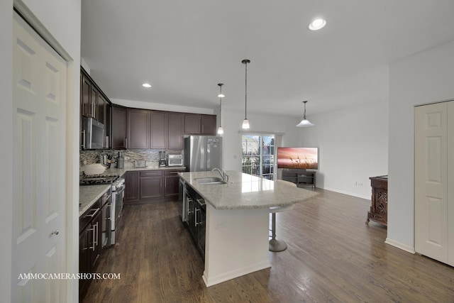 kitchen featuring appliances with stainless steel finishes, pendant lighting, a kitchen island with sink, light stone counters, and dark brown cabinets