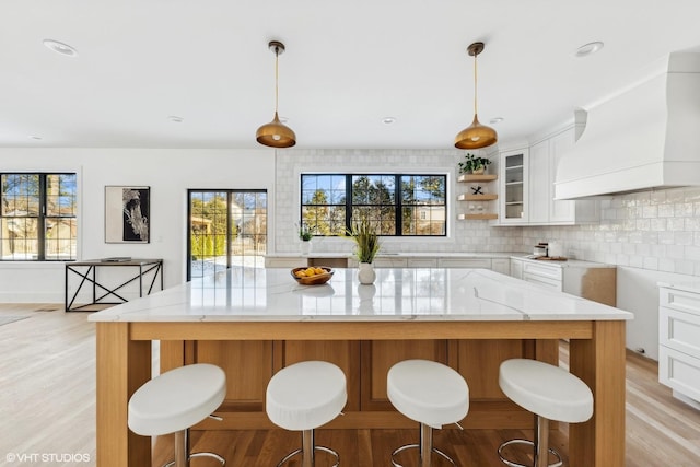 kitchen featuring tasteful backsplash, light stone counters, custom exhaust hood, white cabinetry, and hanging light fixtures