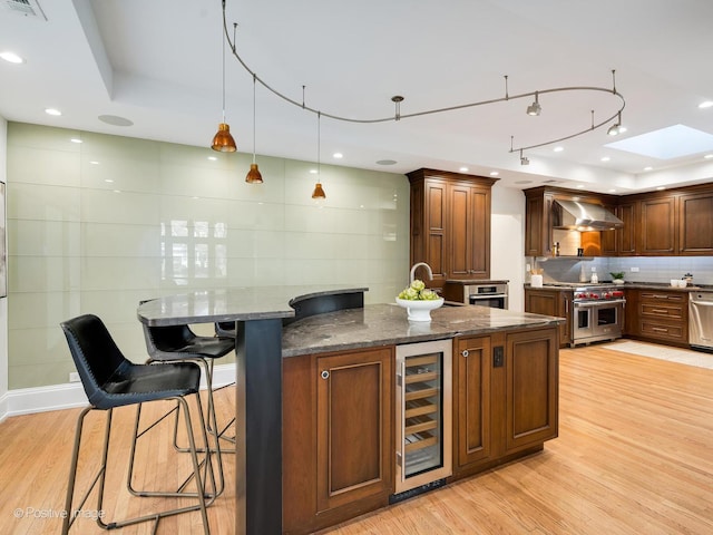 kitchen featuring wall chimney range hood, stainless steel appliances, beverage cooler, dark stone counters, and light wood-type flooring