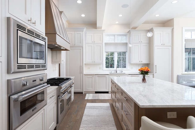 kitchen with beamed ceiling, white cabinets, custom exhaust hood, stainless steel appliances, and light stone countertops